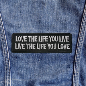 以下が含まれることがあります： 「LOVE THE LIFE YOU LIVE LIVE THE LIFE YOU LOVE」という白い刺繍が施された黒い長方形のパッチが、青いデニム生地に縫い付けられています。パッチはデニムの中央に配置されています。
