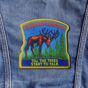 Puede incluir: Un parche con el texto "EVERYBODY GANGSTER" y "TILL THE TREES START TO TALK" sobre un fondo azul. El parche presenta un alce marrón con grandes astas, árboles verdes y un borde amarillo. El parche está cosido a una chaqueta vaquera.
