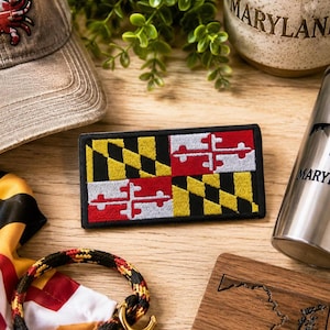 May include: A rectangular embroidered patch featuring the Maryland state flag design with black and gold diagonal stripes in two quadrants and a red and white cross bottony emblem in the other two quadrants, all bordered by a thick black stitched edge.