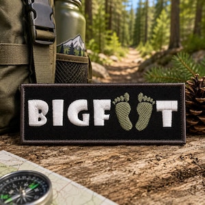 May include: A rectangular black embroidered patch featuring the white text BIGF and T, with two olive green embroidered footprints replacing the letter O. The patch has a dark brown stitched border and is displayed on a textured wooden surface.