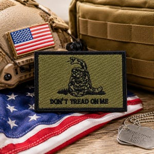 May include: An olive green patch with a black rattlesnake and the text "DON'T TREAD ON ME" sits atop a folded American flag. An American flag patch is on a helmet, and dog tags with the text "FREEDOM ISN'T FREE" are visible.