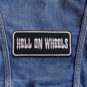 May include: A black rectangular patch with the words "HELL ON WHEELS" embroidered in white gothic lettering. The patch has a white border and is set against a blue denim background.