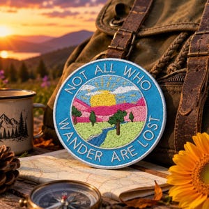 May include: A circular embroidered patch featuring a landscape scene with a yellow sun, pink mountains, green fields, and a blue river. The text "NOT ALL WHO WANDER ARE LOST" is stitched in white along the blue border of the patch.