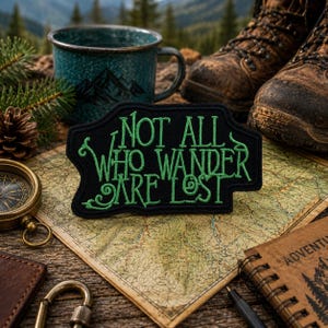 May include: A black embroidered patch with green lettering that reads "NOT ALL WHO WANDER ARE LOST" in a stylized font. The patch has a decorative, irregular border and is positioned on a map, surrounded by hiking gear and outdoor items.