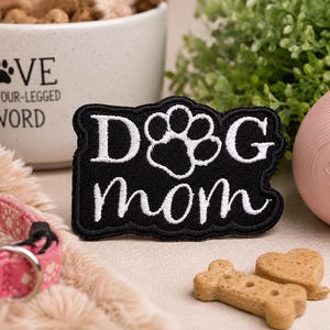 May include: A black embroidered patch with white stitching that reads "DOG mom" with a paw print replacing the letter O. The patch has a scalloped edge and is rectangular in shape with a bold, high-contrast design featuring white text on black.