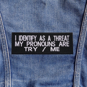 Peut inclure: Un écusson rectangulaire noir avec du texte blanc qui dit "I IDENTIFY AS A THREAT MY PRONOUNS ARE TRY / ME" est cousu sur un tissu en denim bleu. L'écusson est centré et le denim a des coutures visibles.