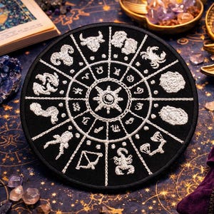 Puede incluir: Un parche circular negro bordado con símbolos astrológicos blancos, dispuestos en un diseño de rueda. El parche está rodeado de cristales, una vela encendida y un libro abierto. El fondo es una superficie azul oscuro con un patrón celestial.