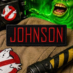 May include: A black patch with the name "JOHNSON" embroidered in red. The patch is on a tan uniform, with a Ghostbusters logo and a proton pack visible. A green, glowing ghost-like figure is in the background.