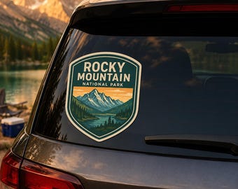 Könnte beinhalten: Ein schildförmiger Aufkleber auf einer Autoscheibe mit dem Text ROCKY MOUNTAIN NATIONAL PARK. Die Grafik zeigt eine Landschaft mit einem blauen See, grünen Kiefern und schneebedeckten Bergen unter einem sanften orange-gelben Himmel.