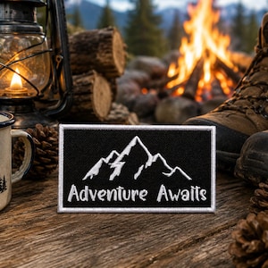 May include: A black rectangular patch with white mountain graphics and the words "Adventure Awaits." The patch is set on a wooden surface, with a lit lantern, a campfire, hiking boots, and a mug in the background. The theme is outdoor adventure.