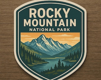 Könnte beinhalten: Ein schildförmiger Aufkleber mit einer malerischen Illustration der Rocky Mountains. Das Design umfasst einen See, immergrüne Bäume und schneebedeckte Berge unter einem orange-gelben Himmel. Der Text "ROCKY MOUNTAIN NATIONAL PARK" ist in Weiß dargestellt.