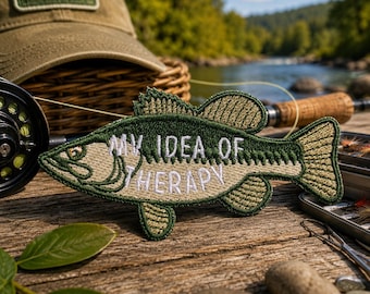 May include: A fish-shaped embroidered patch featuring a dark green body with tan accents and the white stitched text "MY IDEA OF THERAPY". The patch displays detailed fin and scale textures, with a prominent eye and a distinct tail fin design.