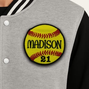 Peut inclure: Gros plan d'une veste de baseball grise et noire avec un écusson de softball jaune. L'écusson porte le nom "MADISON" et le numéro "21" en lettres noires. L'écusson de softball est bordé de noir et a des coutures rouges.