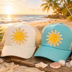 Puede incluir: Dos gorras tipo trucker, una color crema y otra azul claro, cada una con un parche de sol amarillo con borde violeta claro en el panel frontal. Ambas gorras cuentan con una parte trasera de malla y una visera curva en el dise&ntilde;o.