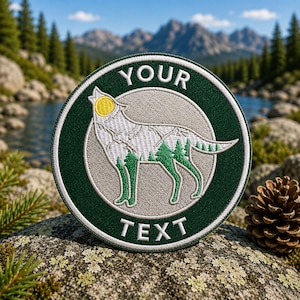 Puede incluir: Un parche bordado circular con un borde verde oscuro con el texto blanco YOUR en la parte superior y TEXT en la inferior. El centro muestra la silueta de un lobo gris relleno con un paisaje forestal verde y un sol amarillo sobre una luna gris.