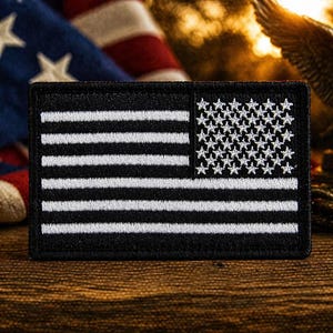 Può includere: Una toppa ricamata in bianco e nero con il disegno della bandiera americana. La toppa ha stelle e strisce bianche su sfondo nero. La toppa rettangolare è esposta su una superficie di legno.