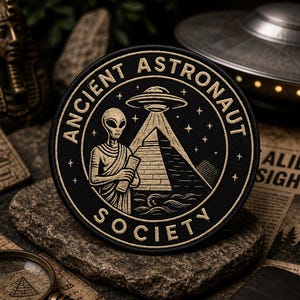 Op de afbeelding: Een zwart en goudkleurig geborduurd embleem met de tekst "Ancient Astronaut Society" rond een alien, een piramide en een UFO. De ronde patch ligt op een grijs stenen oppervlak.