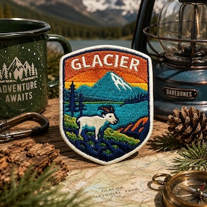 Pode incluir: Patch bordado com uma paisagem de montanha e a palavra "GLACIER" no topo. O patch mostra uma montanha, um lago e uma cabra branca. Uma caneca verde com "ADVENTURE AWAITS" é visível, juntamente com uma lanterna e um mapa.