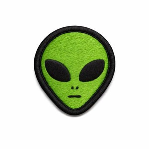 May include: Embroidered patch featuring a green alien face with large black oval eyes and a small mouth. The alien head is outlined in black, creating a distinct border. The patch is likely for clothing or accessories.