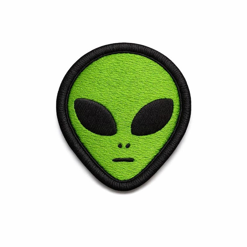 May include: Embroidered patch featuring a green alien face with large black oval eyes and a small mouth. The alien head is outlined in black, creating a distinct border. The patch is likely for clothing or accessories.