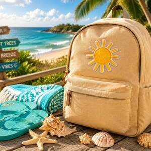 Puede incluir: Una mochila de lona beige con un parche de sol amarillo en el bolsillo delantero. Junto a la mochila hay chanclas turquesas, una toalla turquesa y blanca doblada con estampado, una estrella de mar y tres conchas sobre madera.