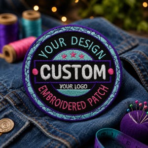 Op de afbeelding: Een ronde, geborduurde patch met de woorden "YOUR DESIGN CUSTOM EMBROIDERED PATCH YOUR LOGO" in wit, roze en teal. De patch ligt op een spijkerjasje, met spoelen garen en een speldenkussen op de achtergrond.