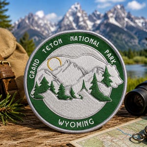 Puede incluir: Un parche bordado circular que presenta la silueta de un oso blanco rellena con una cadena montañosa, pinos verdes y un sol amarillo. El borde verde oscuro muestra el texto Grand Teton National Park y Wyoming en letras blancas.