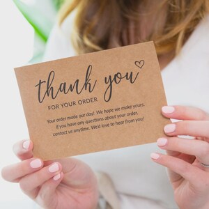 May include: A kraft paper thank you card with the words "thank you" in elegant script, a small heart, and additional text. The card is held by a person with light pink nail polish.