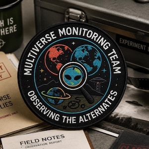 Op de afbeelding: Een ronde zwarte geborduurde patch met de tekst MULTIVERSE MONITORING TEAM en OBSERVING THE ALTERNATES. Het ontwerp bevat drie planeten, een vergrootglas met een alien-gezicht, een kleine UFO en sterren op een donkere achtergrond.