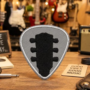 May include: A gray guitar pick-shaped patch with a black guitar headstock design. The patch is on a wooden surface with a small chalkboard sign that reads "LIVE LOVE PLAY". A small card reads "GOOD VIBES GOOD MUSIC".