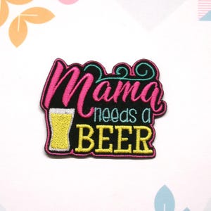 May include: Embroidered iron-on patch with black background, pink text that says "Mama needs a" and yellow text that says "BEER". A yellow beer glass graphic is to the left of the text.