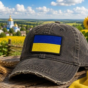 May include: A distressed black baseball cap with a rectangular patch featuring the Ukrainian flag: blue over yellow. The cap sits on a wooden surface with wheat stalks and a sunflower in the background. A scenic view of a field and church is visible.