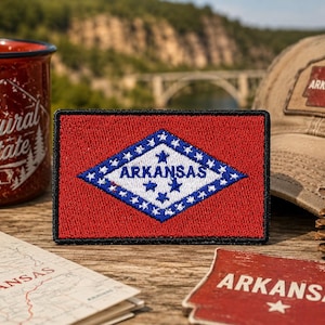 May include: A rectangular embroidered patch featuring the Arkansas state flag design. The patch has a red background with a central white diamond bordered in blue, containing the word ARKANSAS in blue text surrounded by blue stars.