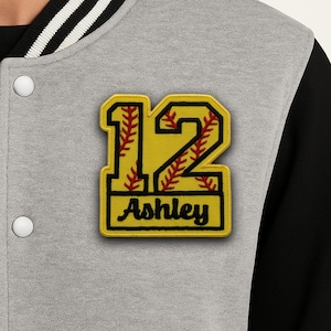 May include: A grey letterman jacket with a yellow baseball-themed patch. The patch features the number 12, red stitching, and the name "Ashley" in black lettering. The jacket has black sleeves and white and black striped trim.
