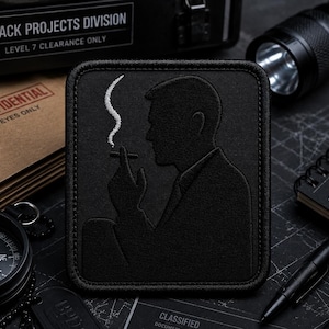 Op de afbeelding: Een rechthoekige zwarte geborduurde patch met het silhouet van een persoon in een pak die een sigaret vasthoudt, met een enkele witte geborduurde rookpluim die omhoog stijgt. De patch heeft een dikke gestikte rand en een matte zwarte stofafwerking.