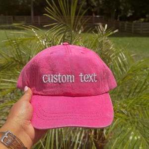 May include: A vibrant pink baseball cap with the embroidered text "custom text" in white. The cap features a curved brim and a classic six-panel design. The hat is made of a soft, textured fabric.