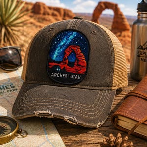 Op de afbeelding: Een verweerde antracietgrijze truckerpet met een rechthoekige patch met een kleurrijke geborduurde afbeelding van een rode rotsboog onder een sterrenhemel, met de tekst ARCHES UTAH in het wit onderaan de patch.