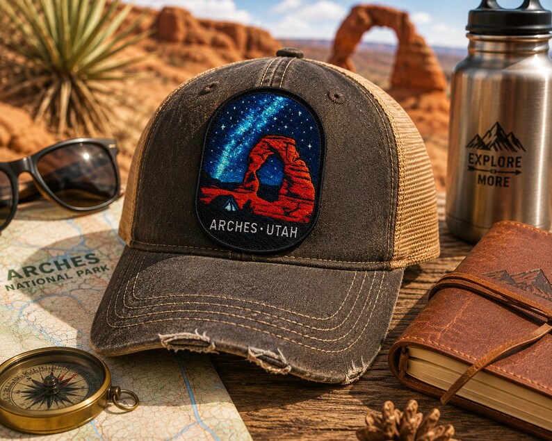 Op de afbeelding: Een verweerde antracietgrijze truckerpet met een rechthoekige patch met een kleurrijke geborduurde afbeelding van een rode rotsboog onder een sterrenhemel, met de tekst ARCHES UTAH in het wit onderaan de patch.