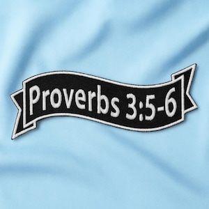Puede incluir: Un parche negro en forma de banner con texto blanco que dice "Proverbios 3:5-6" está centrado en una tela azul claro. El parche tiene un borde blanco y una forma decorativa en cada extremo. La tela tiene una apariencia sutil y texturizada.