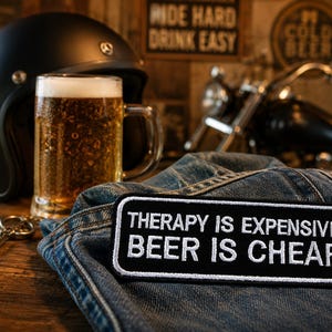 May include: A black motorcycle helmet, a full beer mug, and a denim jacket with a patch that reads "THERAPY IS EXPENSIVE BEER IS CHEAP" are displayed on a wooden surface. A motorcycle is in the background.