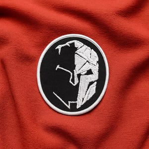 May include: A circular embroidered patch with a black and white Spartan helmet design. The helmet is white, set against a black background, with a white border. The patch is on a textured, red-orange fabric.