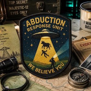 May include: A shield-shaped embroidered patch featuring a dark blue night sky with yellow stars and text reading ABDUCTION RESPONSE UNIT and WE BELIEVE YOU. A flying saucer emits a yellow beam lifting a cow and a person above a green landscape.