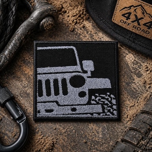 May include: Black square patch featuring a gray embroidered Jeep vehicle design. The patch is surrounded by a black carabiner, a black hat with a 4x4 Off Road patch, and a black rope.
