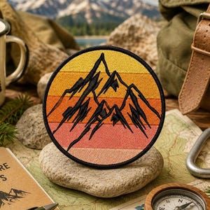 Puede incluir: Un parche bordado circular que presenta la silueta de una cordillera negra estilizada sobre un fondo de franjas horizontales en tonos degradados de amarillo, naranja y rosa, todo encerrado dentro de un borde negro cosido.