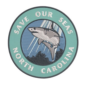 May include: Embroidered patch with a teal border and the text "SAVE OUR SEAS NORTH CAROLINA". The patch features a detailed illustration of a shark leaping out of the water, with stylized rays of light and a dark blue background.