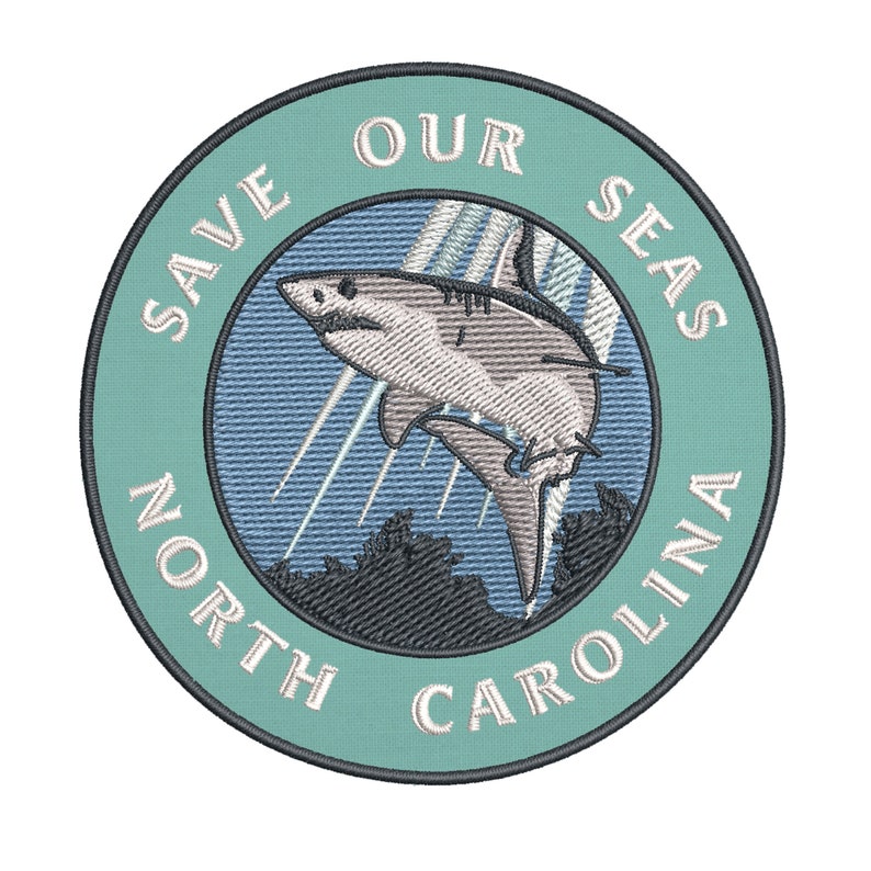 May include: Embroidered patch with a teal border and the text "SAVE OUR SEAS NORTH CAROLINA". The patch features a detailed illustration of a shark leaping out of the water, with stylized rays of light and a dark blue background.