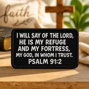 May include: Black rectangular patch with white text: "I WILL SAY OF THE LORD, HE IS MY REFUGE AND MY FORTRESS, MY GOD, IN WHOM I TRUST. PSALM 91:2." A wooden cross and a book are in the background.