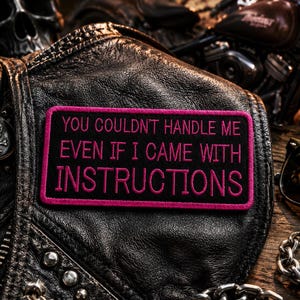 May include: A black leather jacket with a patch that reads "YOU COULDN'T HANDLE ME EVEN IF I CAME WITH INSTRUCTIONS" in pink lettering. The jacket has a skull and crossbones button and is near a motorcycle and sunglasses.