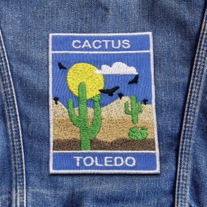 May include: Embroidered patch featuring a desert scene with cacti, a yellow sun, and the text "CACTUS" and "TOLEDO" on a blue background. The patch is rectangular with a white border and is attached to a denim jacket.