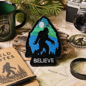 Könnte beinhalten: Ein pfeilspitzenförmiger bestickter Aufnäher mit der schwarzen Silhouette einer Kreatur vor einer blau-grünen Landschaft mit weißem Mond und dem Wort BELIEVE in weißer Schrift am unteren Rand, umgeben von einer Tasse, einem Notizbuch und einer Kamera.